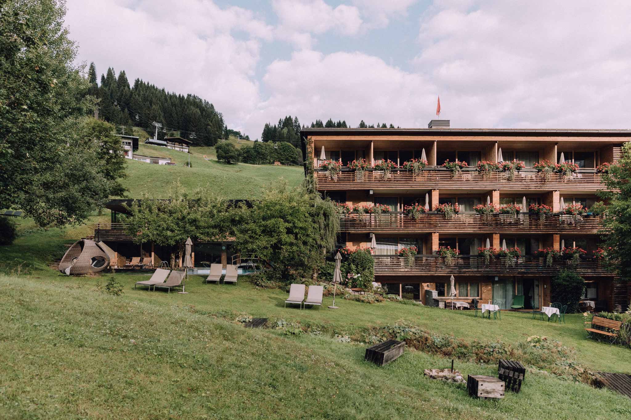 Blick aus dem Garten auf Haus Chesa mit Alpin Spa, grüner Wiese, Bach und Außenpool