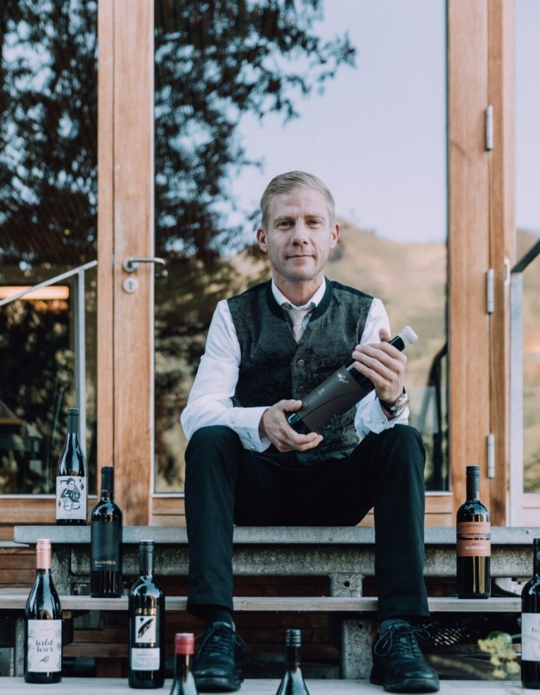 Attila sitzt vor dem Wintergarten umringt von Weinflaschen