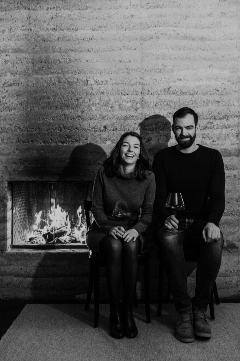 Magdalena und David Kessler sitzen lachend am Kamin mit Rotwein